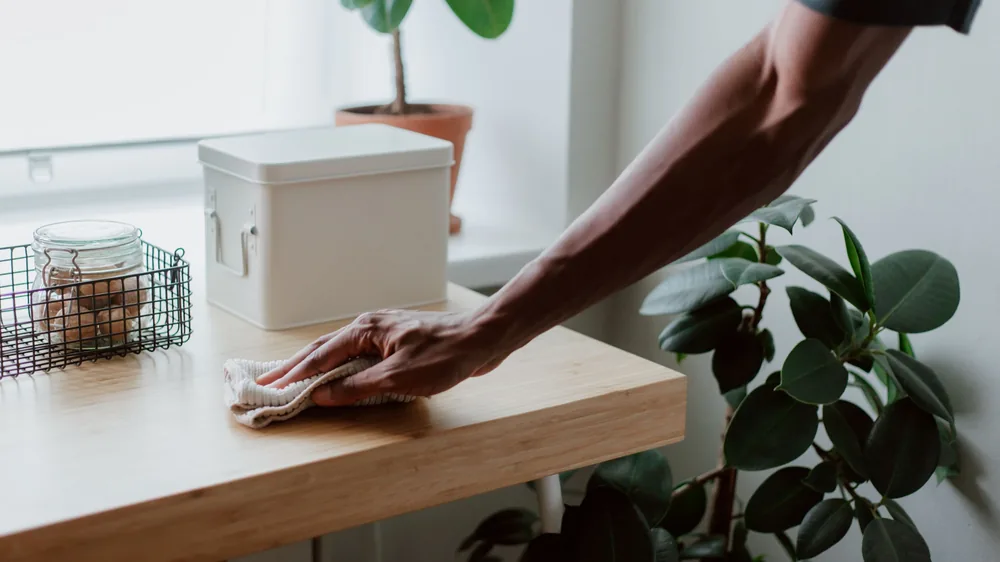Hand wiping a bamboo desk with a cloth beside potted houseplants