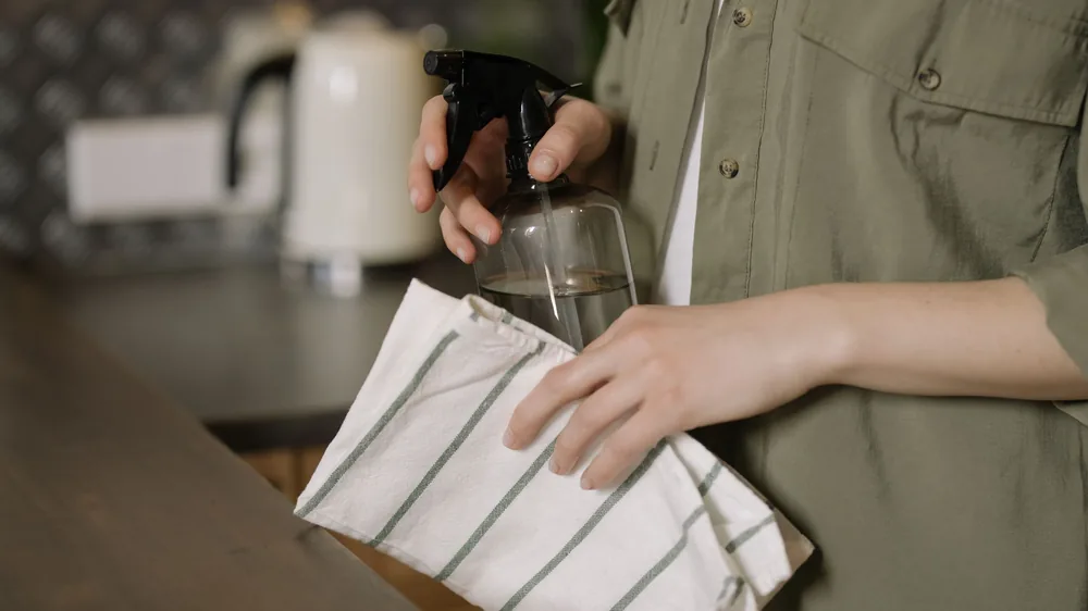Hands holding a glass spray bottle and striped cloth at a kitchen worktop