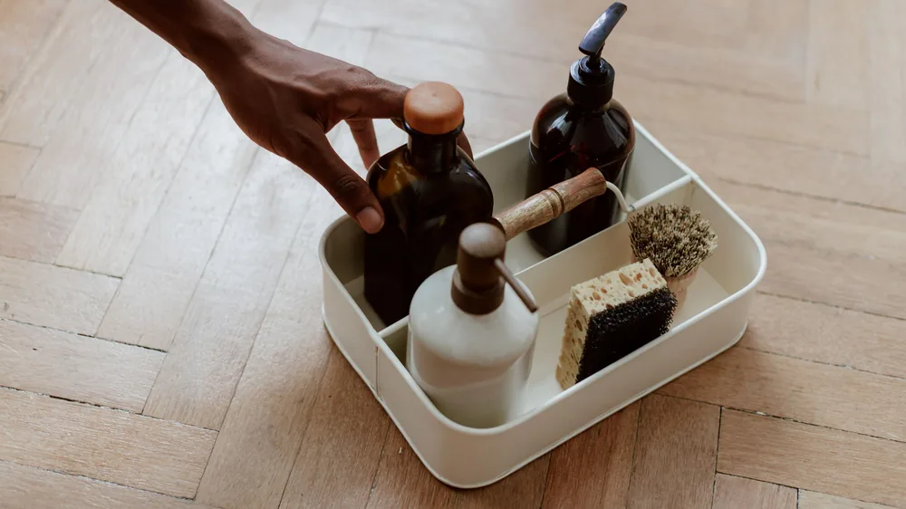Hand reaching into a cleaning caddy of bottles and brushes on parquet