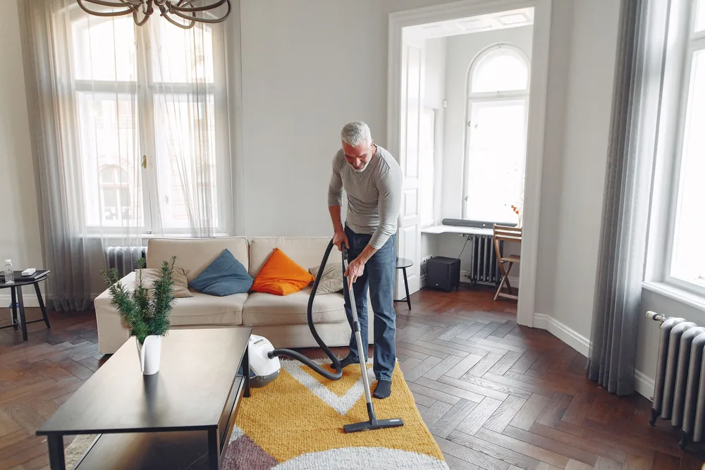 Grey-haired man vacuuming a yellow rug in a bright living room