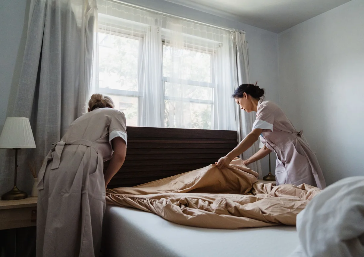 Two cleaners in uniform making a bed by a sunlit window